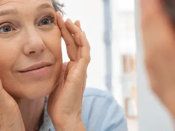 Stock photograph of a model: a gray-haired woman touches the skin beside her eyes as she looks into a mirror