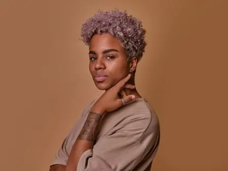 Stock photograph of a model: a person with short lilac curls and a tattooed forearm poses against a tan backdrop