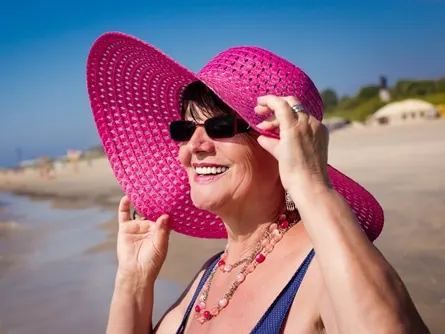 Stock photograph of a model: a smiling older woman in a pink sun hat and sunglasses on a beach