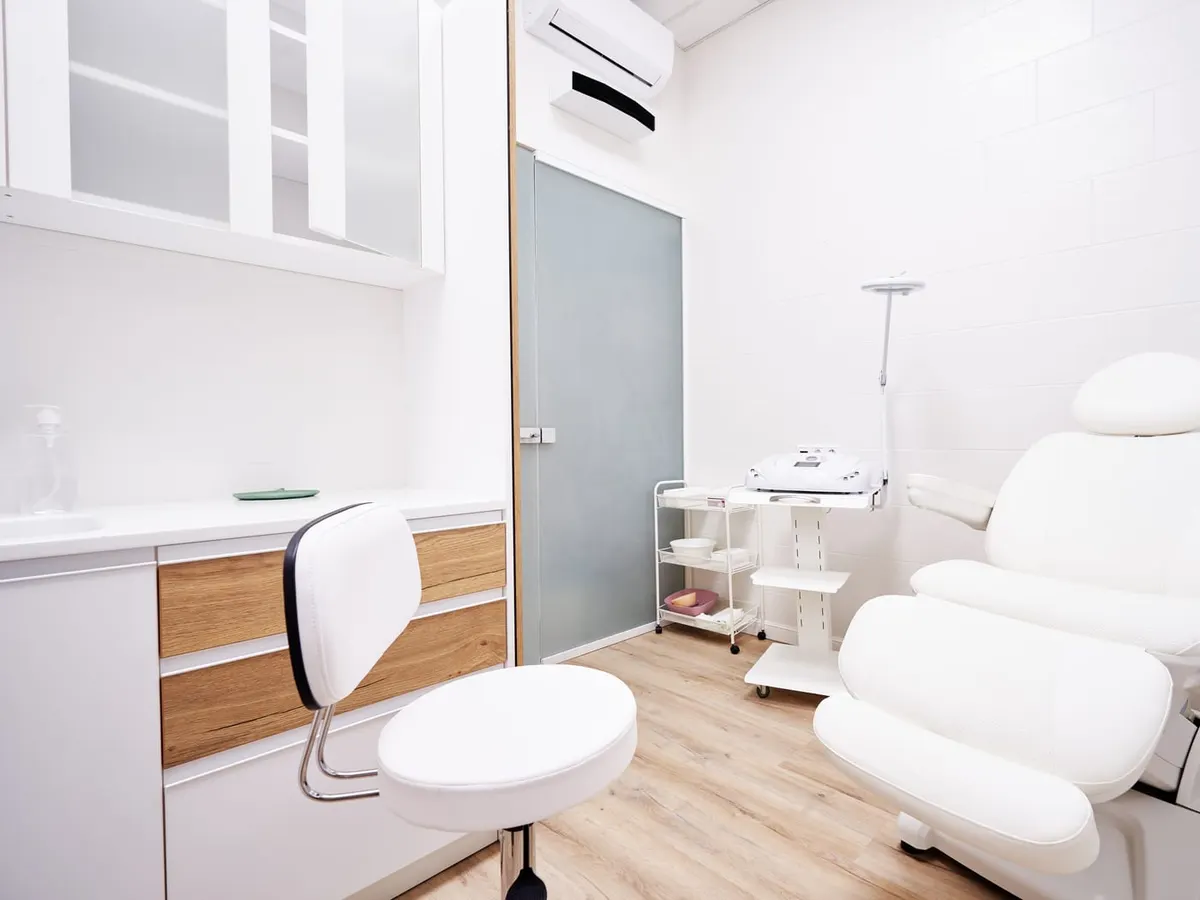 An empty, bright treatment room with a white reclining treatment chair, a stool, a small equipment cart and white cabinets over a wood-look floor