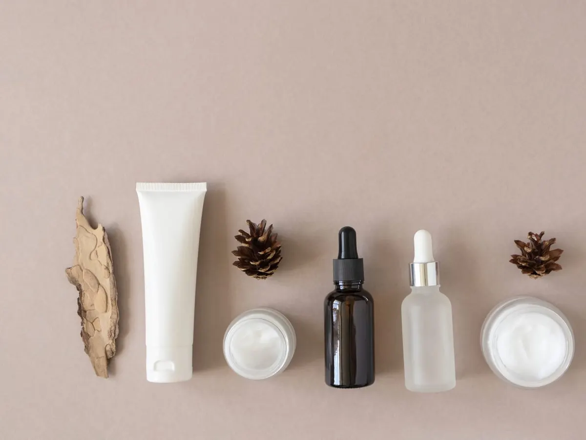 Unlabelled skincare products in a row on a beige surface: a white tube, cream jars and two dropper bottles, between pine cones and pieces of bark