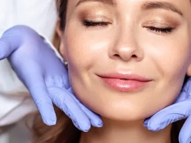Stock photograph of a model: a woman with closed eyes and a faint smile as gloved hands in lavender gloves cradle her jaw, a white-coated clinician behind her.
