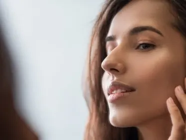 Stock photograph of a model: a woman looking at her reflection in a mirror with her fingertips lightly touching her cheek near her ear.