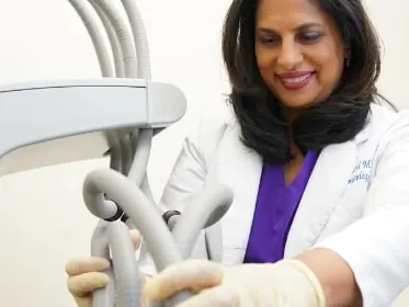 Dr. Nita Patel, smiling in a white coat over purple scrubs, adjusts the hoses of a laser treatment machine in an exam room.