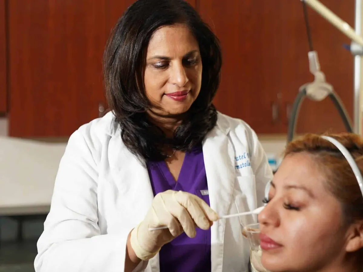 Dr. Nita Patel in a white coat and purple scrubs applies a chemical peel solution with a cotton swab to the face of a woman wearing a white headband.