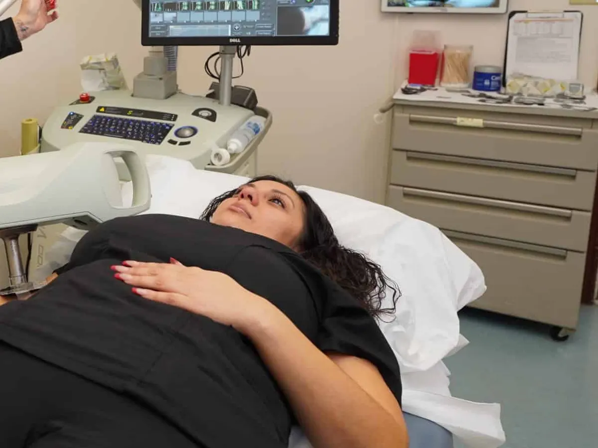A woman in black scrubs lies on a treatment table while a clinician in black positions the arm of a superficial radiation therapy machine over her.