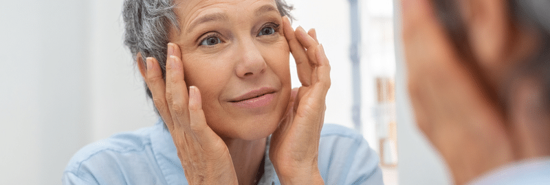 Stock photograph of a model: a gray-haired woman touches the skin beside her eyes as she looks into a mirror