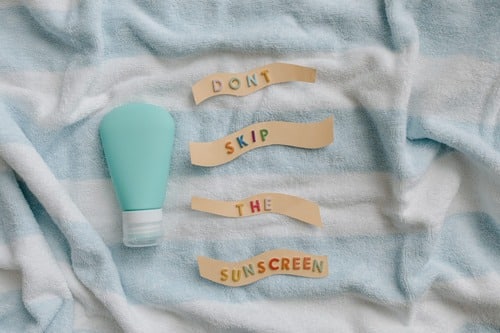 Stock photograph: a teal sunscreen bottle on a striped towel beside paper strips spelling "Don’t skip the sunscreen"