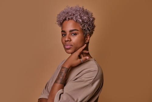 Stock photograph of a model: a person with short lilac curls and a tattooed forearm poses against a tan backdrop