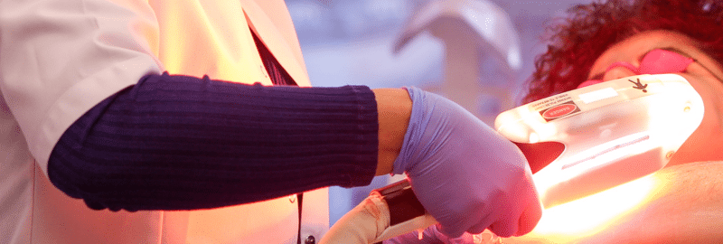 Stock photograph of models: a gloved clinician holds a glowing light-therapy handpiece over a patient wearing eye shields