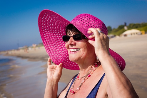 Stock photograph of a model: a smiling older woman in a pink sun hat and sunglasses on a beach