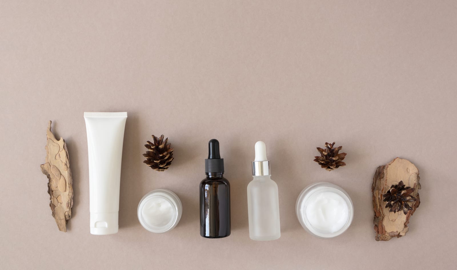 Unlabelled skincare products in a row on a beige surface: a white tube, cream jars and two dropper bottles, between pine cones and pieces of bark