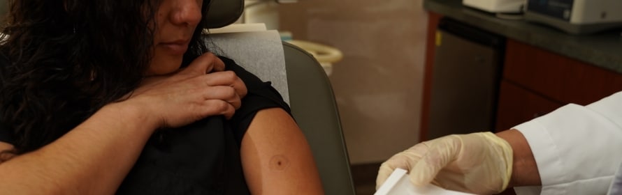 A woman in a black top sits in a dermatology chair while a gloved clinician in a white coat holds paper near her upper arm, which has a small round mole.